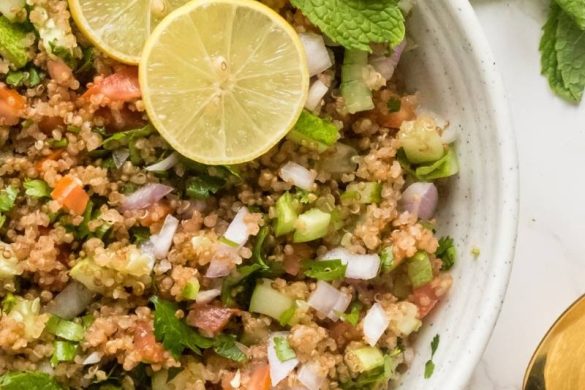 Quinoa tabbouleh in a white serving bowl topped with fresh lemon slices and fresh mint alonmg with golden spoons.