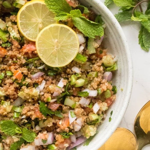 Quinoa tabbouleh in a white serving bowl topped with fresh lemon slices and fresh mint alonmg with golden spoons.
