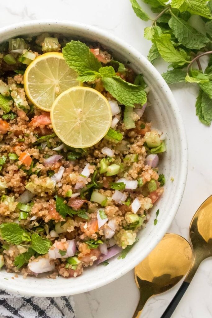 Quinoa tabbouleh in a white serving bowl topped with fresh lemon slices and fresh mint alonmg with golden spoons.