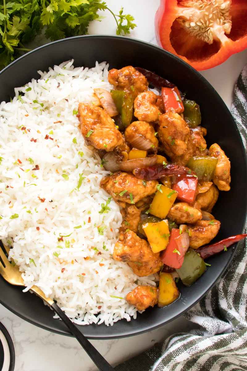 Szechuan Chicken recipe in a black plate with a golden and balck spoon alont with fresh cilantro and red bell pepper