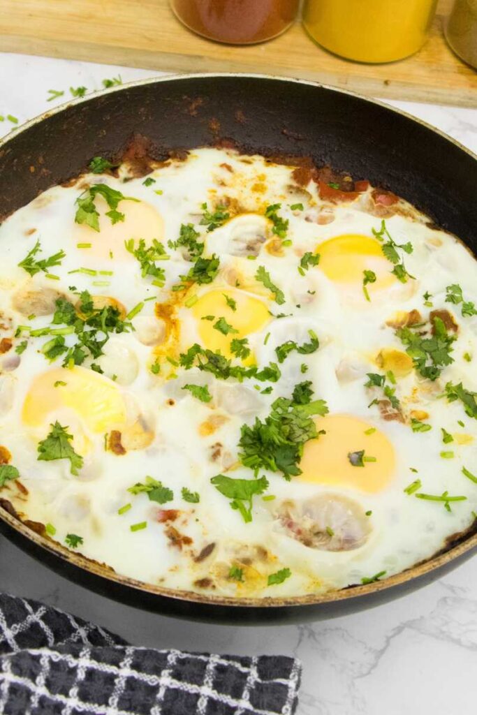 Shakshuka in pan garnished with cilantro nearby with spice jars on the side and showing cooked eggs in a rich middle eastern sauce