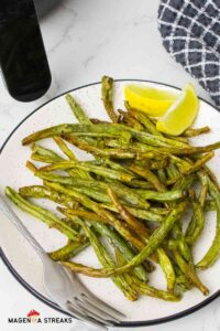 close up to air fryer green beans on a white plate with 2 lemon wedges