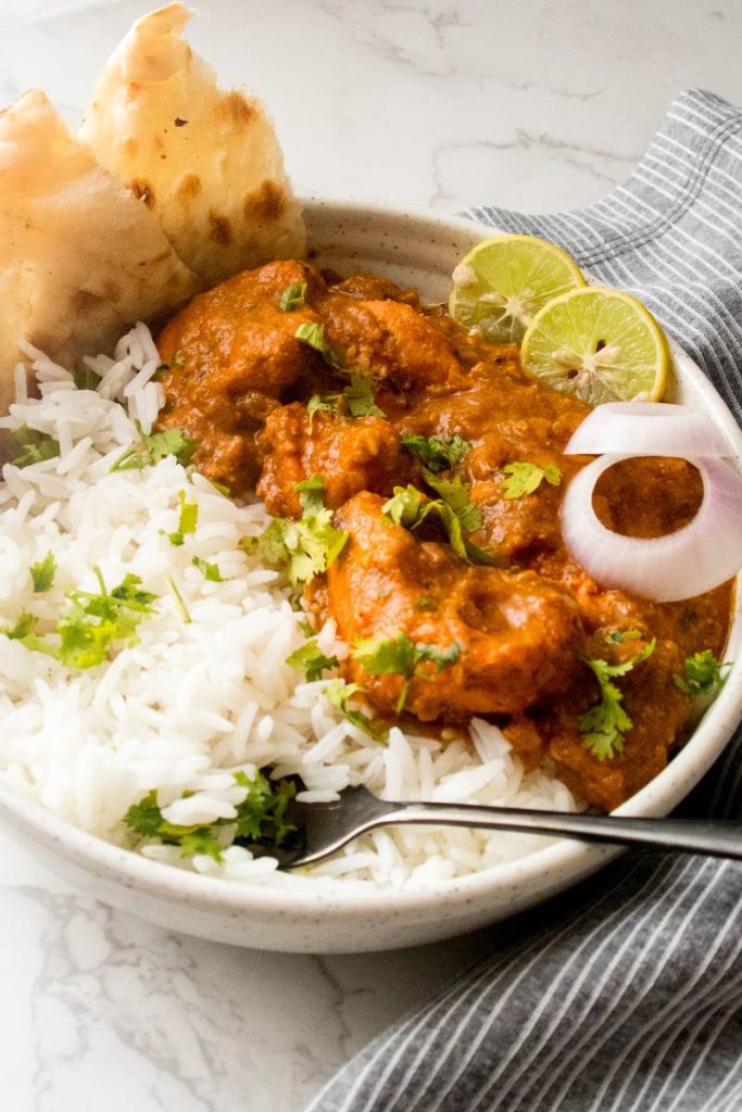 A bowl full of chicken tikka masala over basmati rice, onion rings, lemon slices and with a slice of naan.